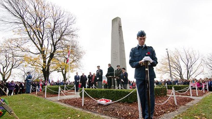 A solemn ceremony at a war memorial with a crowd in the background and trees in autumn.