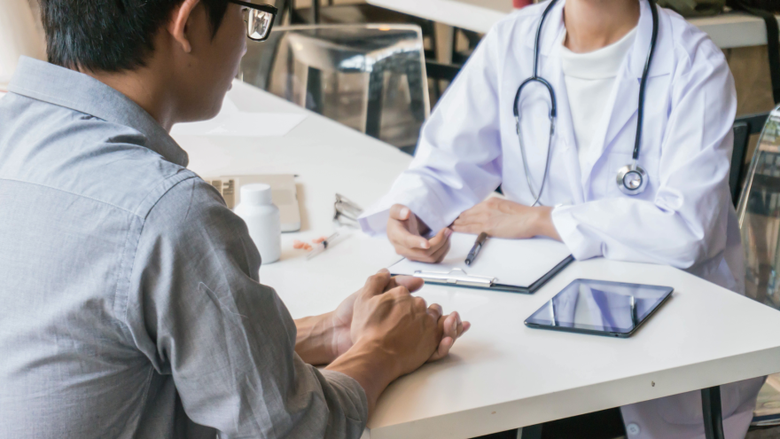 A healthcare professional speaks with a patient at a table, with a tablet and documents present.